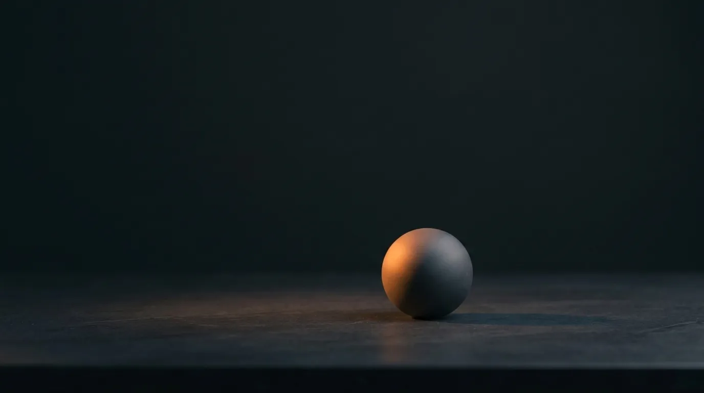 A single matte grey sphere resting on a dark stone surface against a near-black background, lit from one side by a warm orange glow that catches its left edge. Vast empty negative space surrounds it.