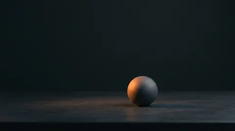 A single matte grey sphere resting on a dark stone surface against a near-black background, lit from one side by a warm orange glow that catches its left edge. Vast empty negative space surrounds it.
